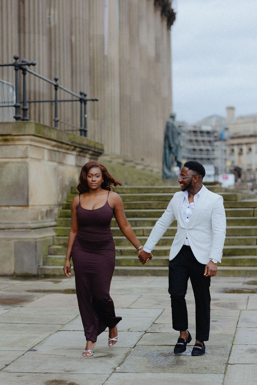 A couple in contrasting outfits dances playfully on stone steps near classical columns.