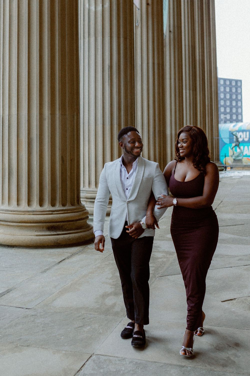 A couple in formal attire walks together between elegant stone columns in an architectural setting.