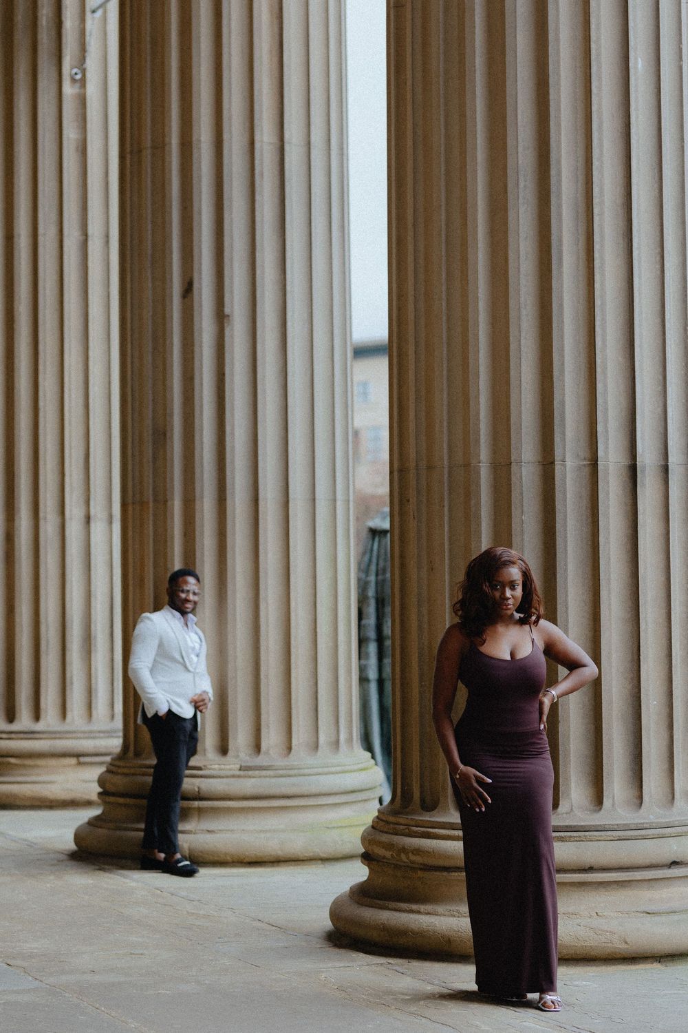 Two people pose separately between classical stone columns in an engagement photo session.