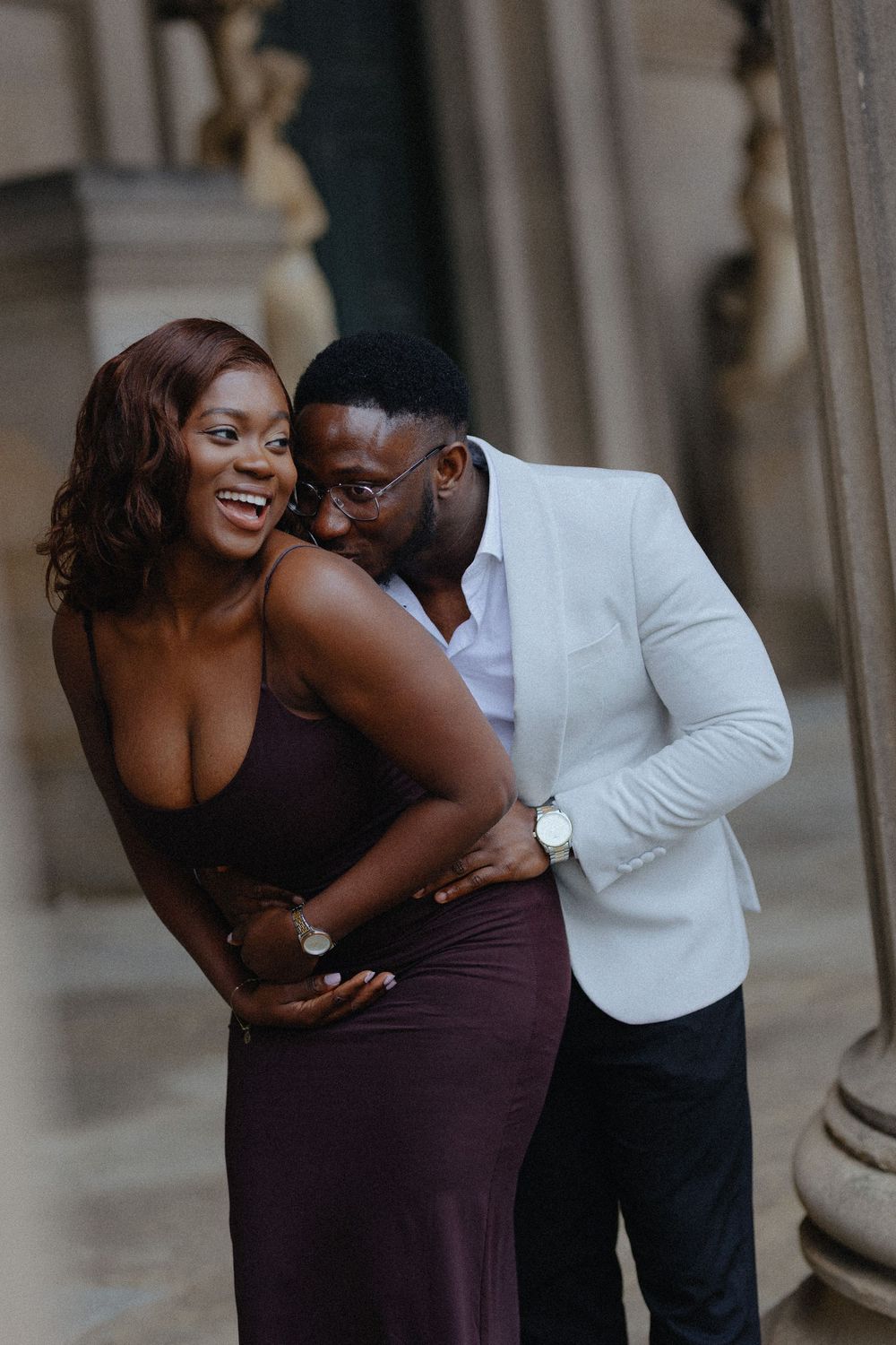 A romantic couple shares intimate moments between elegant stone columns during a photo shoot.