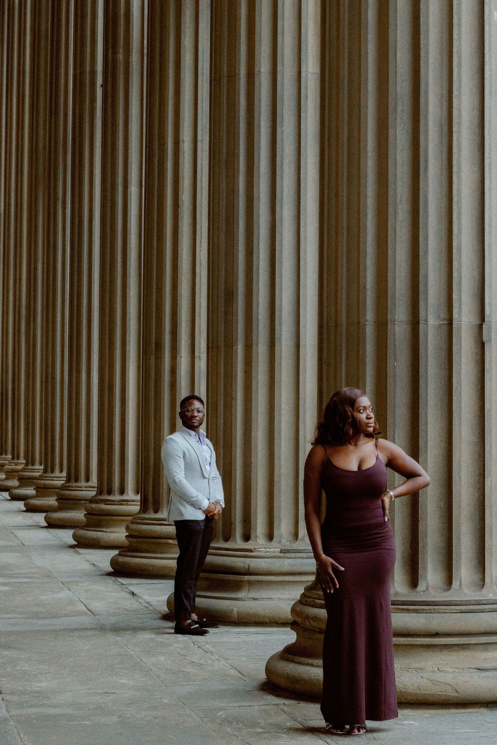 Two people dance between dramatic columns in an architectural setting with strong shadows.
