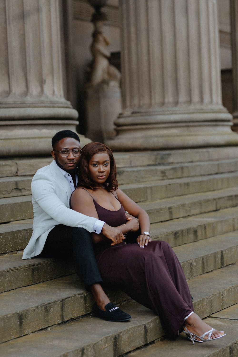 A couple sits together on stone steps near columns in formal attire during an engagement photoshoot.