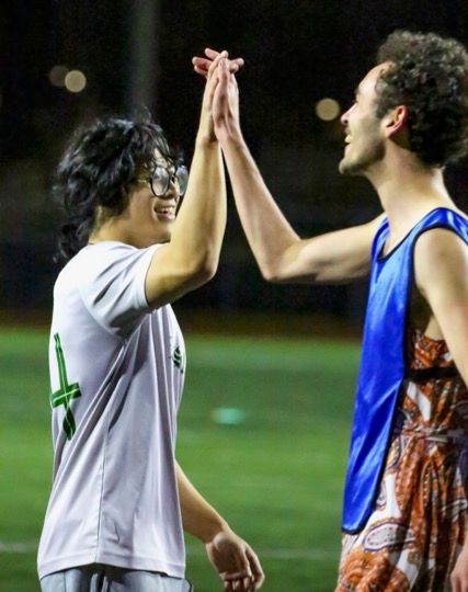 Male Soccer player in white and blue jerseys high-five during a night time soccer match.