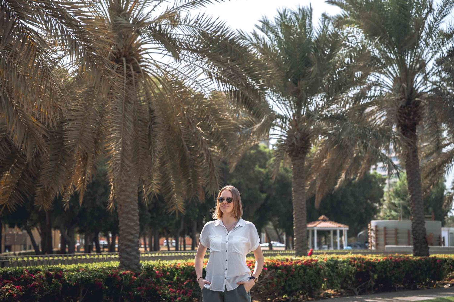 Woman wearing sunglasses and a white shirt stands confidently among palm trees in a sunny Abu Dhabi park.