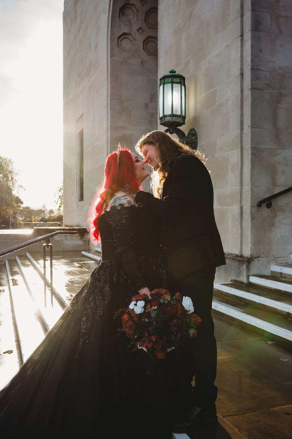 A couple embraces on stone steps at sunset with dramatic sunlight streaming through architectural columns.