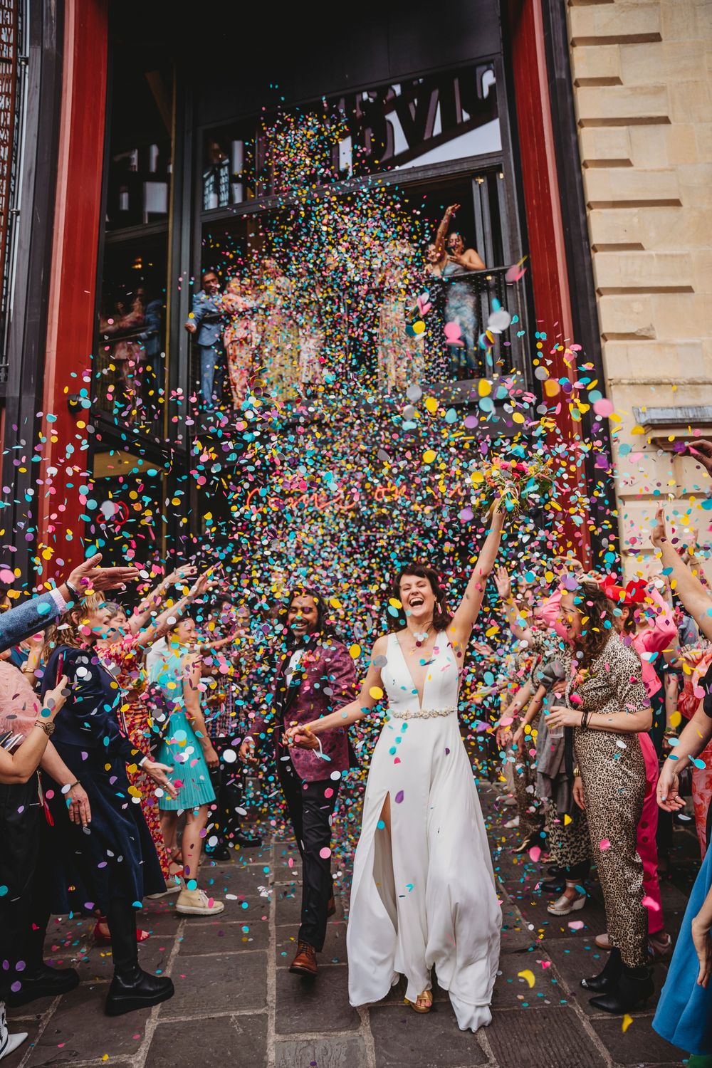 Colorful confetti rains down on a joyous celebration outside a building entrance with a red doorframe.