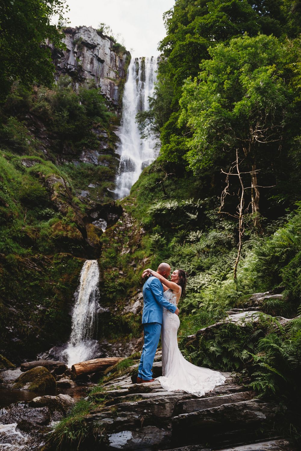 A couple embraces in front of a cascading waterfall surrounded by lush greenery in a romantic outdoor setting.