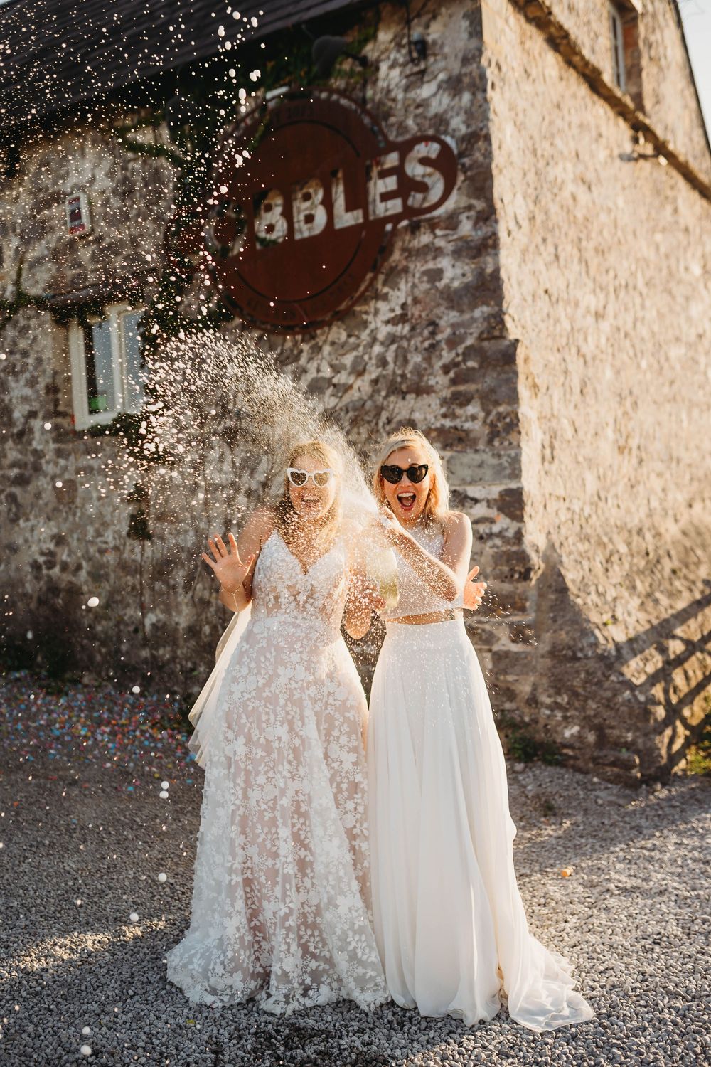 Two people in white wedding dresses pose together with confetti falling around them against a rustic stone wall.
