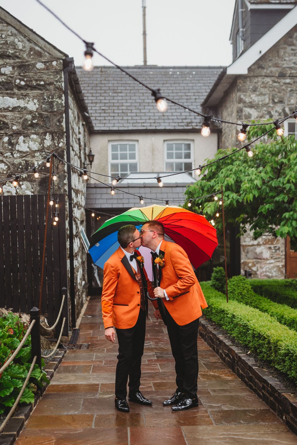 Two people in matching orange jackets share a moment under colorful umbrellas in a rainy garden setting.