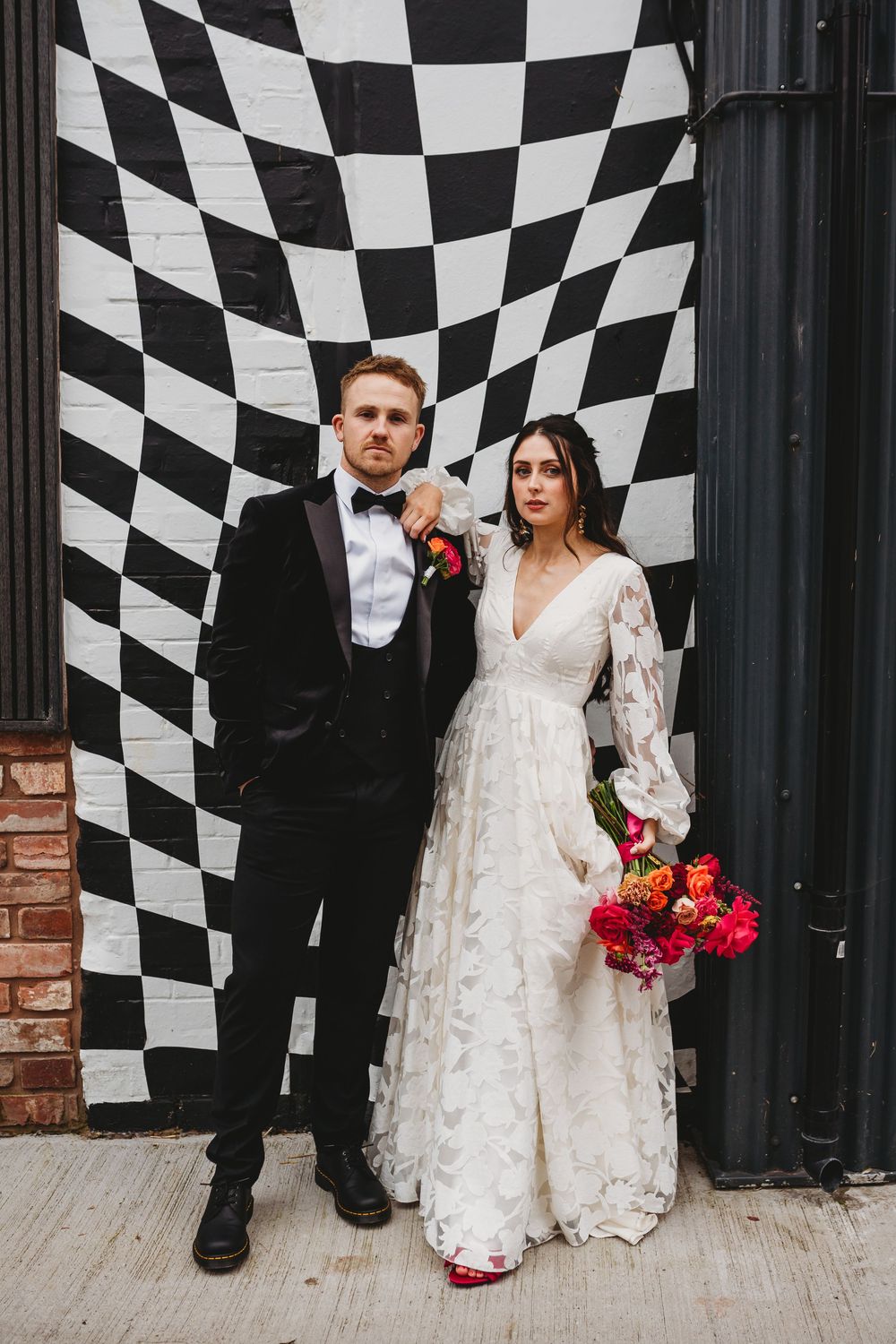 Wedding couple poses against black and white checkered backdrop with red bouquet and white lace dress.