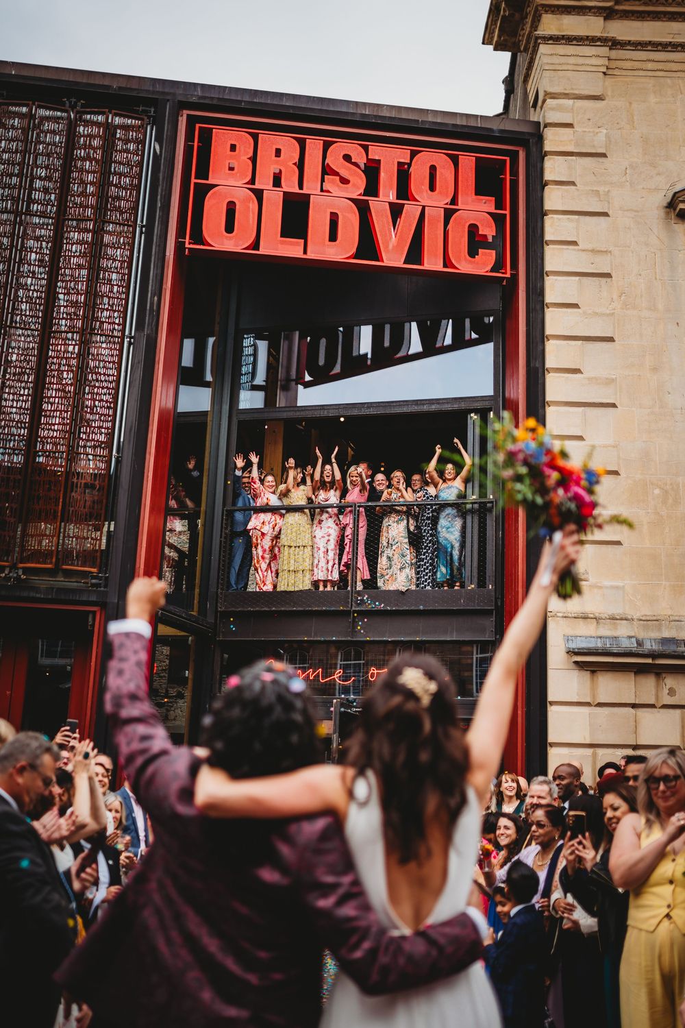 Wedding guests celebrate and raise their hands in joy outside the Bristol Old Vic theater entrance.