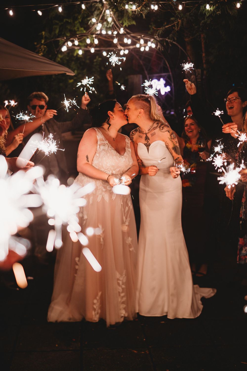 A romantic wedding celebration at night with sparklers glowing while two people in white gowns share a tender moment.