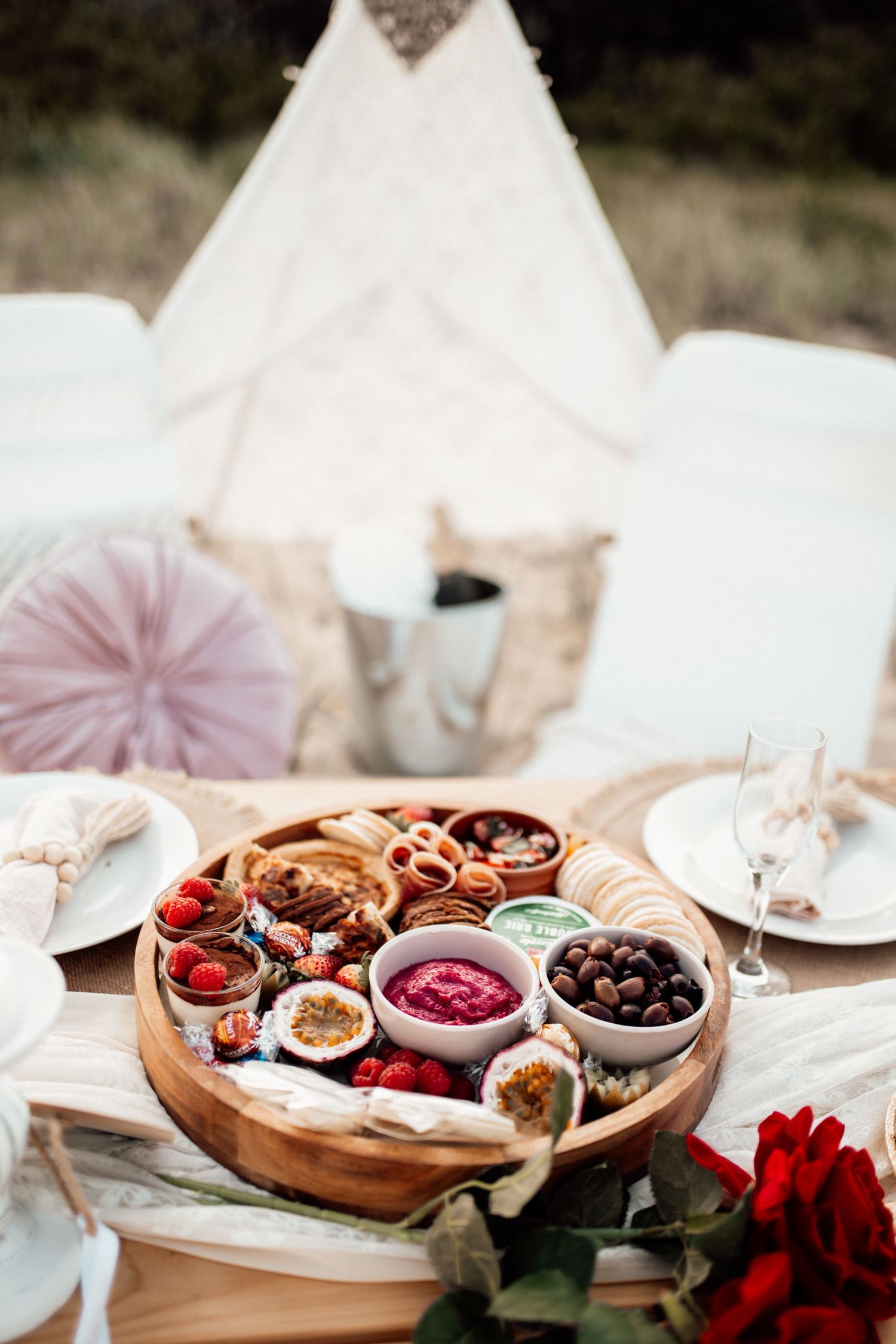 A rustic charcuterie board with dried fruits, jams, and red roses displayed on an elegant white table setting.