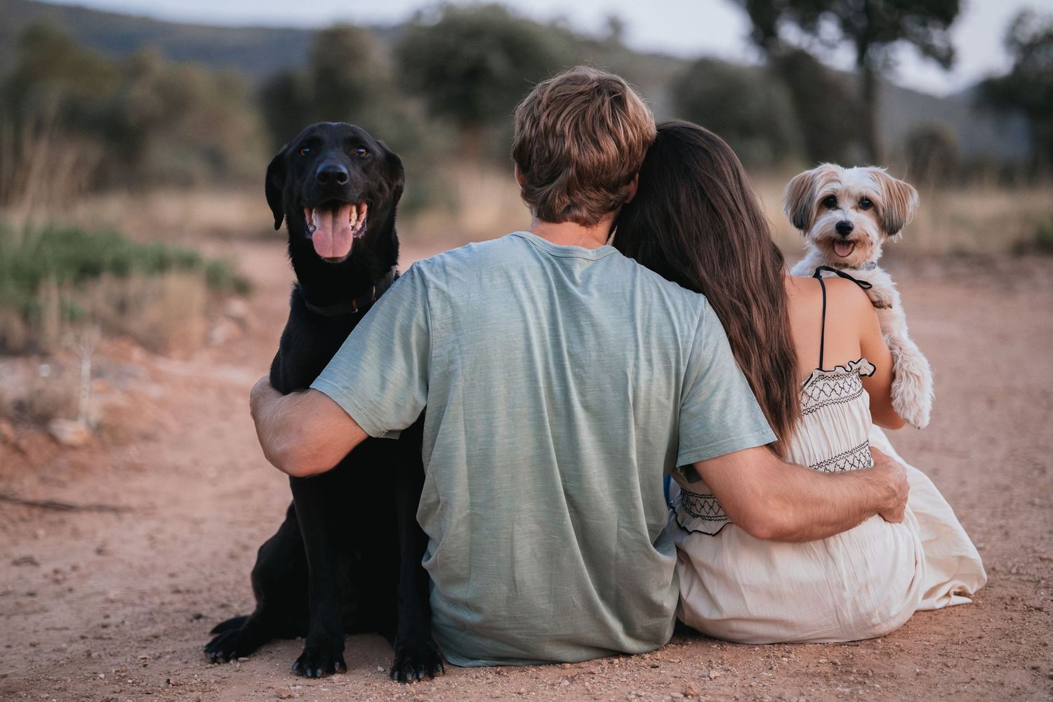 Couple sitting on dirt path with small white dog in natural outdoor setting.