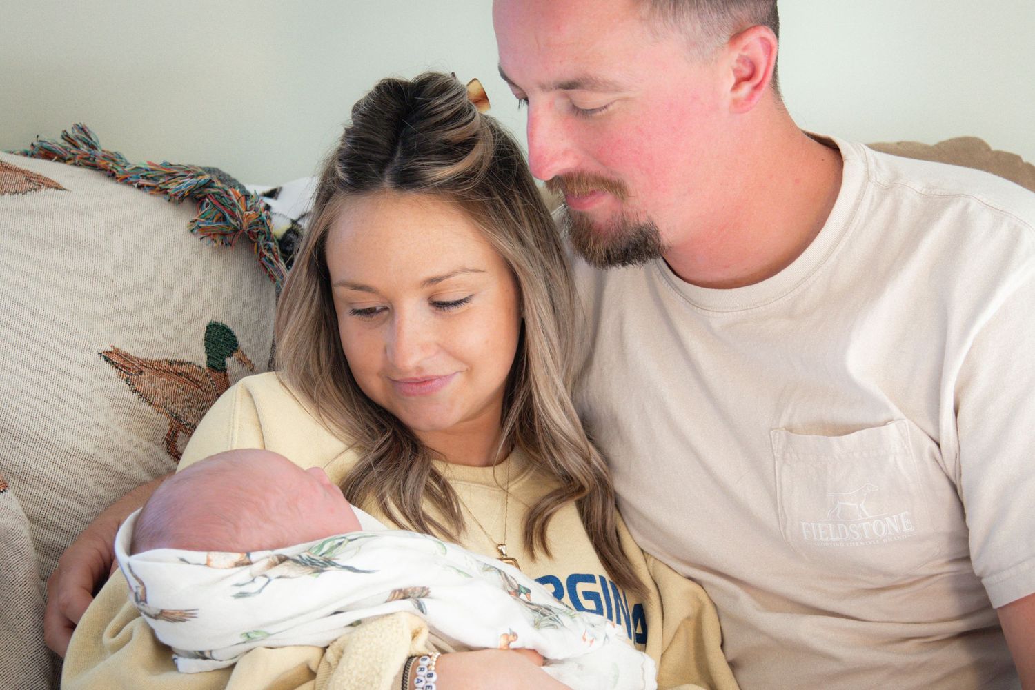 A couple resting together on a couch while holding their newborn baby wrapped in swaddling blanket.