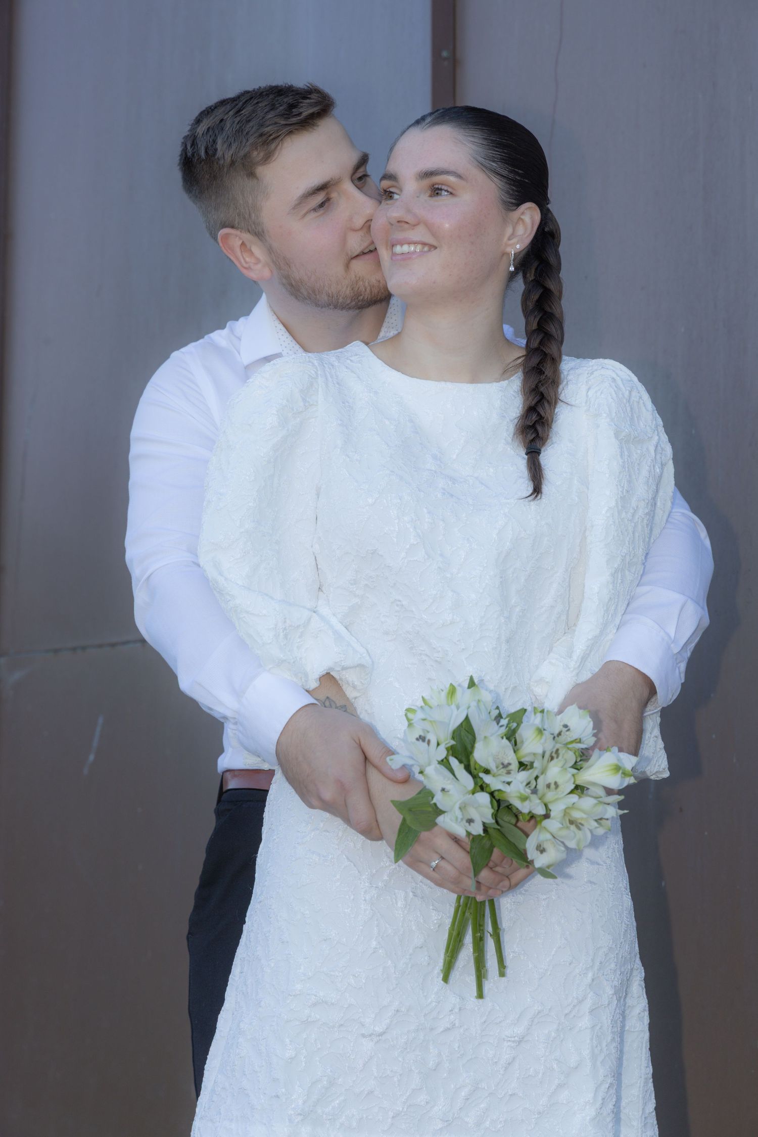 Groom kissing bride on cheek as she smiles holding white flower bouquet in elegant white wedding dress.