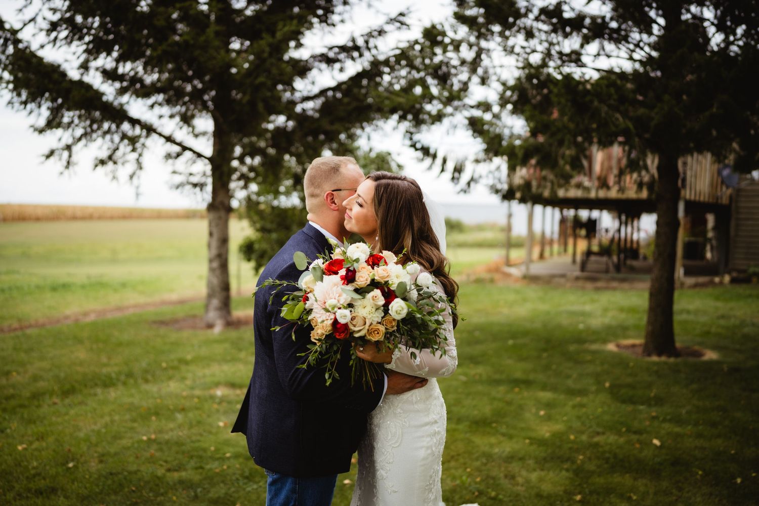 Wedding couple embraces among tall trees during an outdoor fall ceremony near a rustic venue.