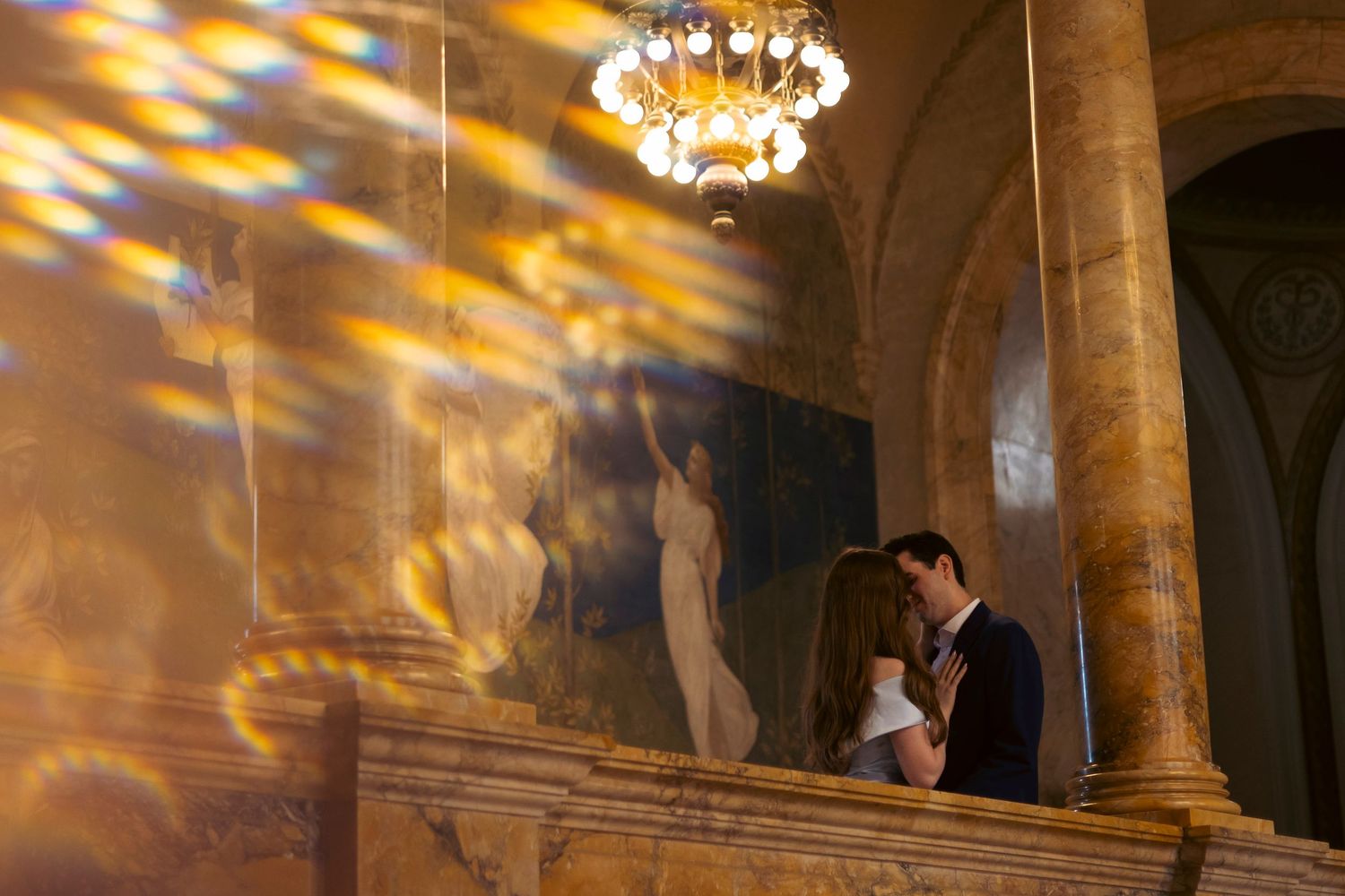 A couple shares an intimate moment beneath a crystal chandelier casting warm reflections across marble columns and walls.