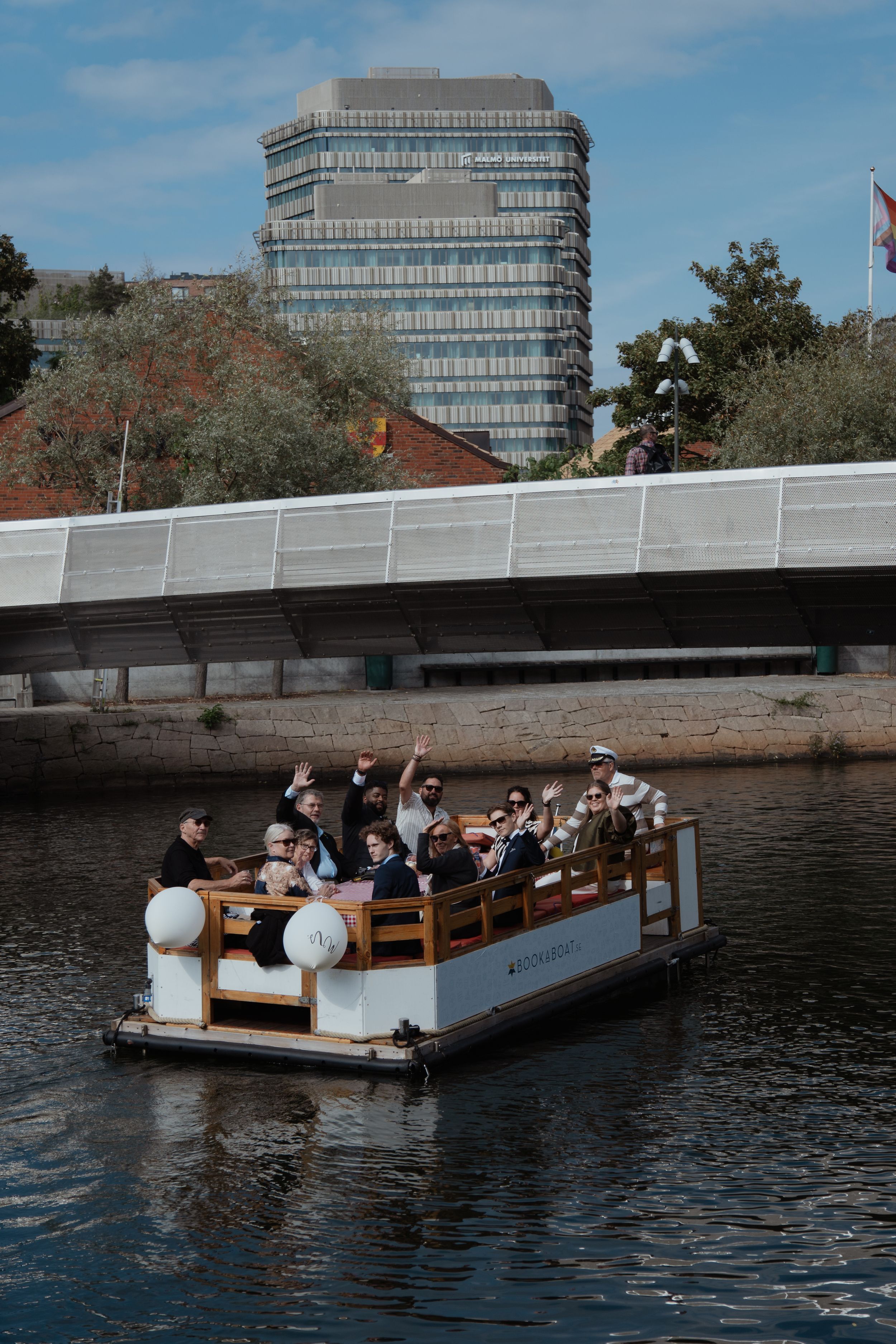 A group enjoys a scenic boat ride on a small wooden platform vessel floating along an urban waterway on a sunny day.