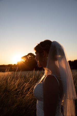 A bride in a flowing veil stands silhouetted against a golden sunset in a field of tall grass.