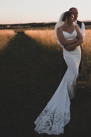 Couple in wedding attire stands embracing in golden field at sunset with lace dress details visible in the warm light.