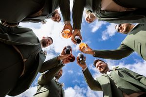 Groomsmen form a circle and look down at the camera against a blue sky with white clouds.