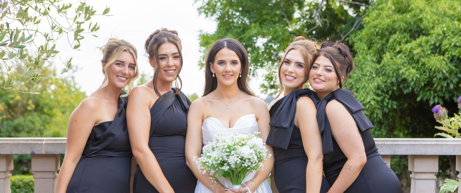 Bridesmaids wearing navy blue dresses pose together outdoors near elegant white columns and greenery.