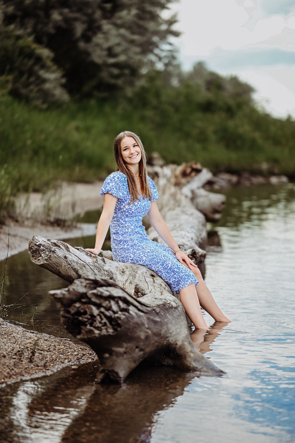 A woman in a blue floral dress sits on a fallen log by the water's edge on a sunny summer day.