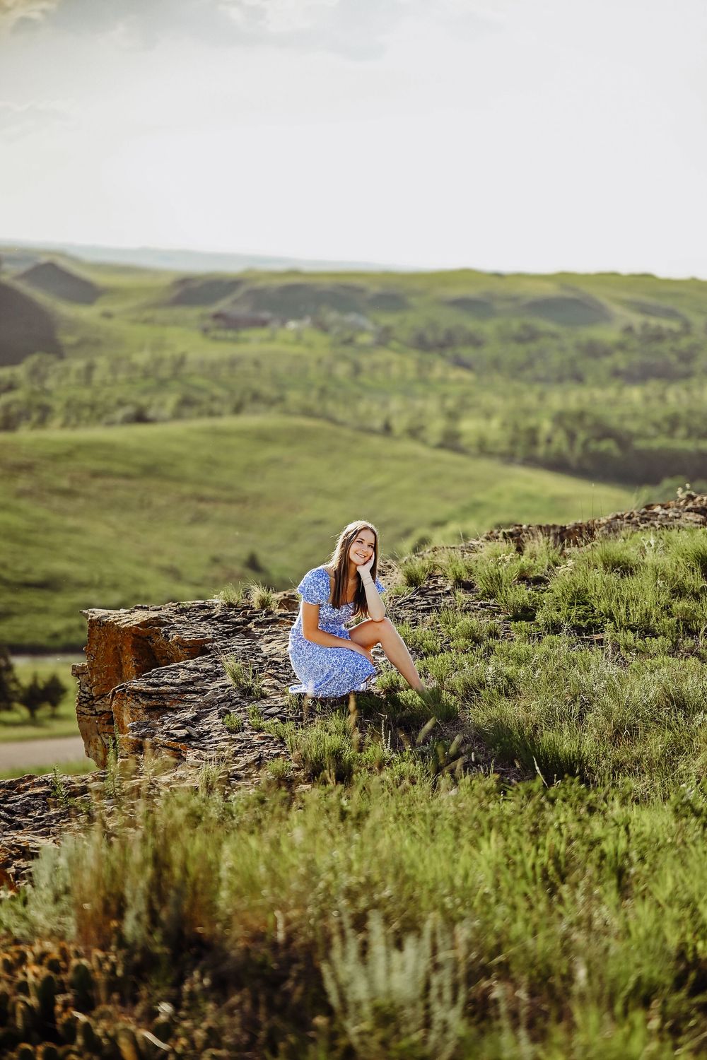 A person in a blue dress sits on a stone wall overlooking rolling green hills and scenic countryside landscape.