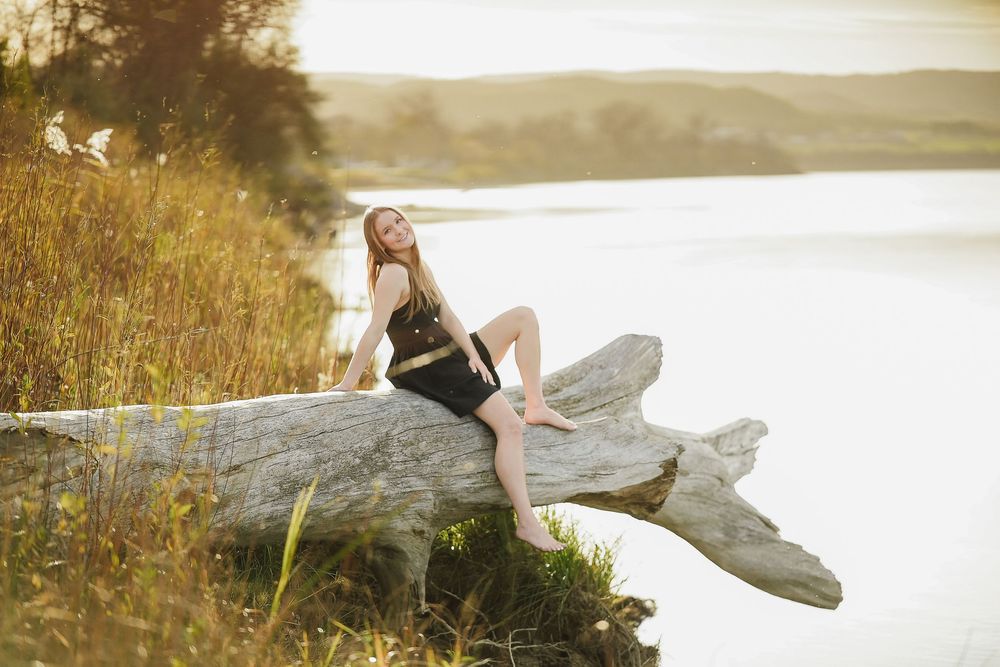 A person in a flowing white dress poses gracefully by a lake at sunset creating an ethereal scene.