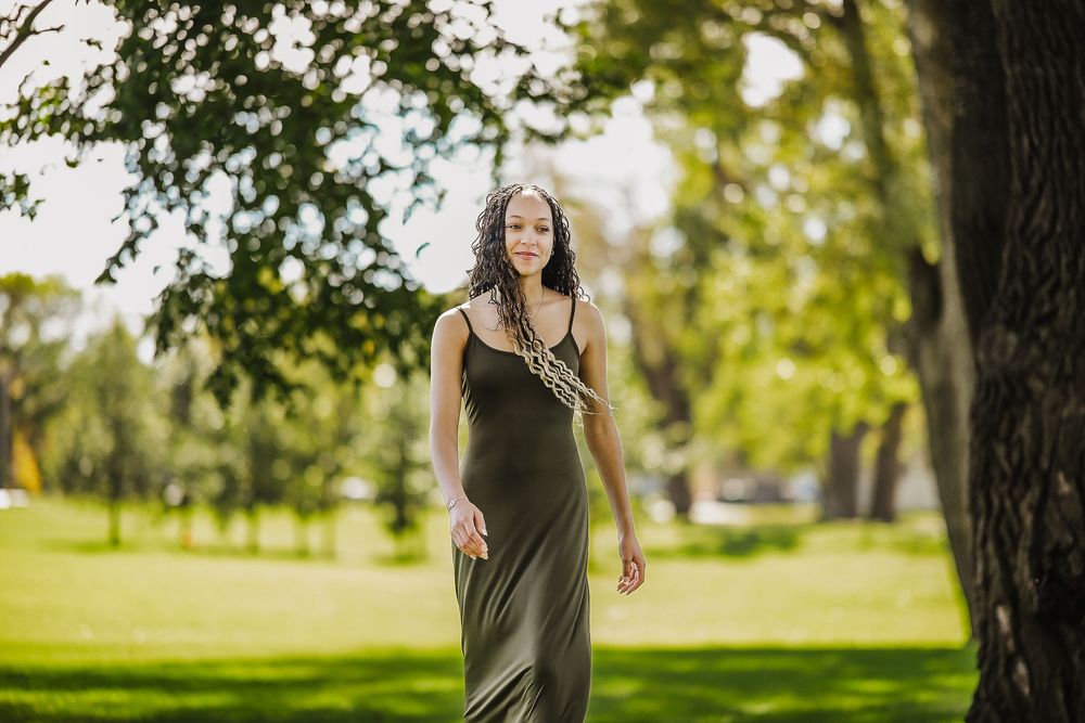 Woman in olive green dress poses outdoors in natural sunlit setting with trees and grass in background.