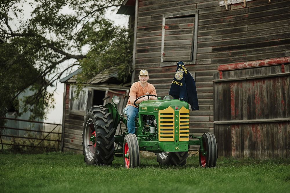 People gather near an old weathered barn with farm equipment visible in the background.