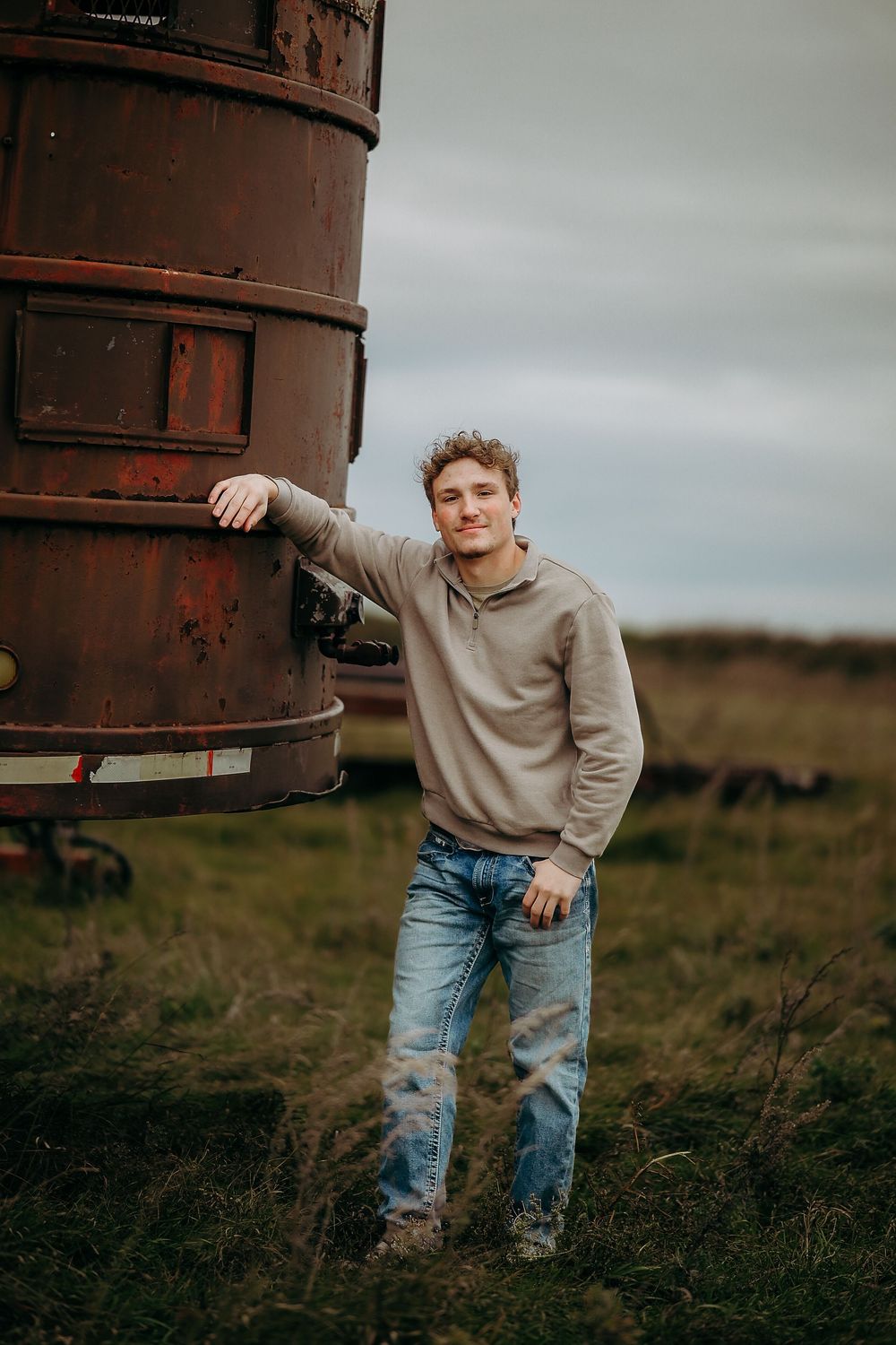 Person in beige sweater and jeans poses playfully against rustic metal silo in rural countryside setting.