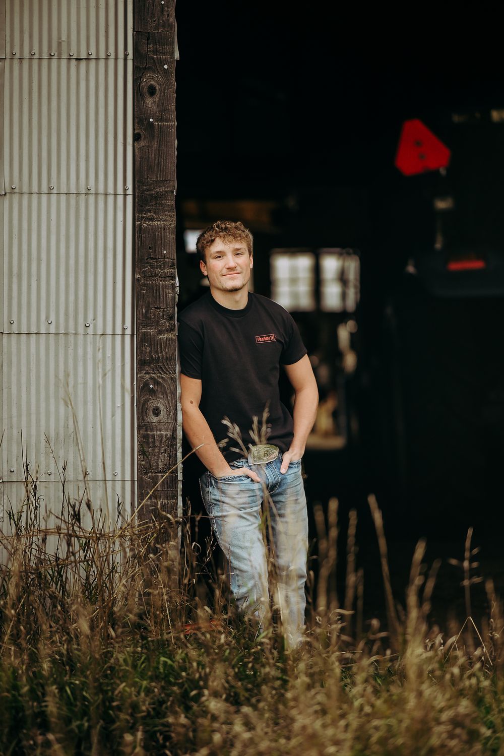 A person stands in tall grass against a rustic wooden column wearing jeans and a black shirt in moody lighting.