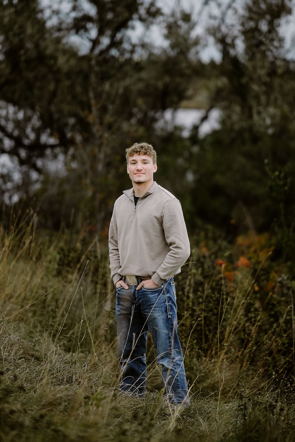 A person in a beige sweater and jeans poses in a field of tall grass during an autumn outdoor photoshoot.