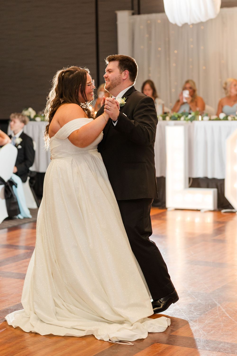 A graceful wedding couple dances together in an elegant ballroom with soft lighting and classic white decor.