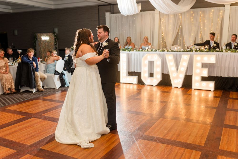 Couple shares their first dance on a wood parquet floor with illuminated LOVE letters and wedding decor in background.