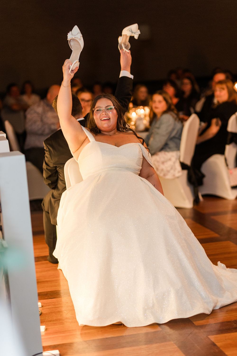 A bride in a strapless white ballgown dances joyfully on a wooden dance floor at her wedding reception.