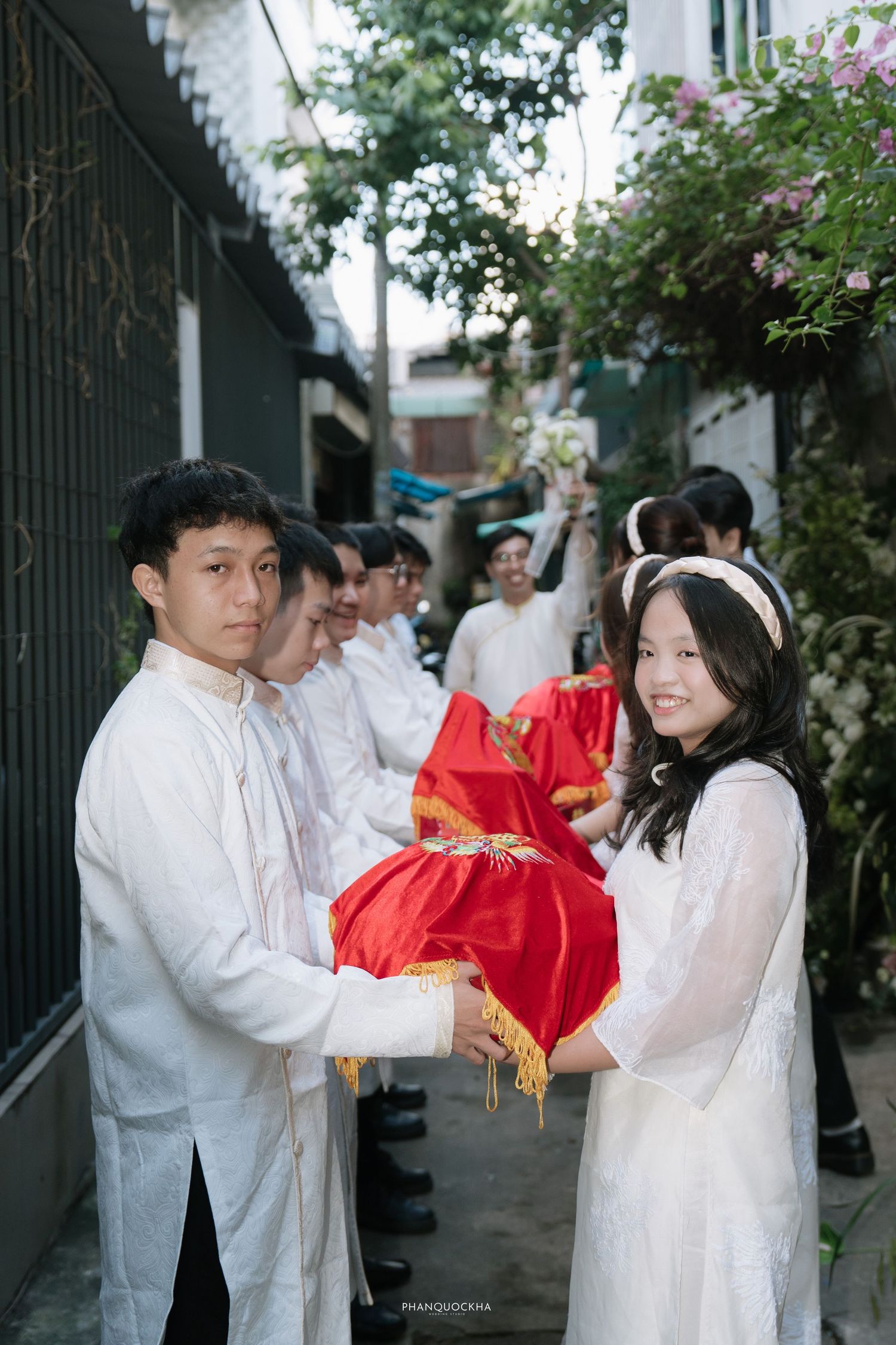 Wedding party in white ao dai passes red ceremonial gift boxes during Vietnamese engagement ceremony.