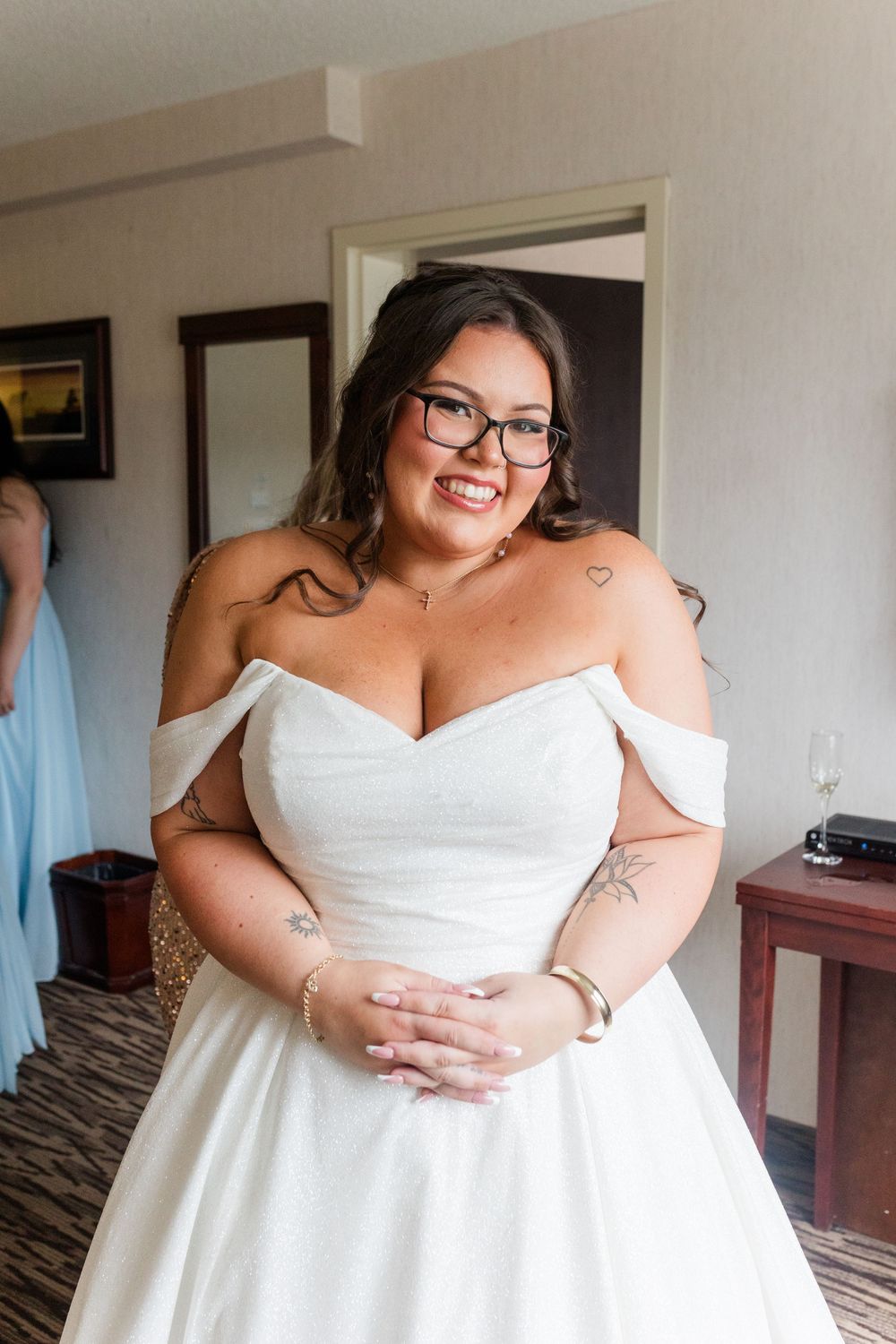 A person in a white strapless wedding dress with glasses adjusts their gown in a hotel room.