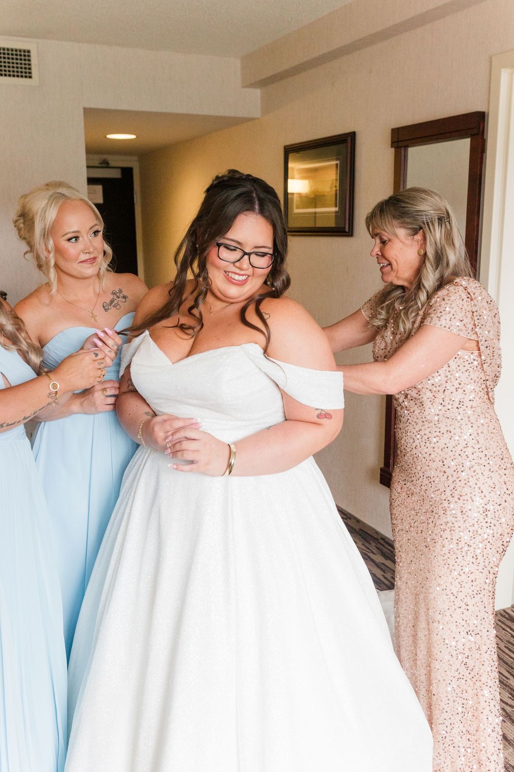 Bridesmaids in pastel dresses help adjust a bride's off-shoulder white wedding gown in a hotel room.