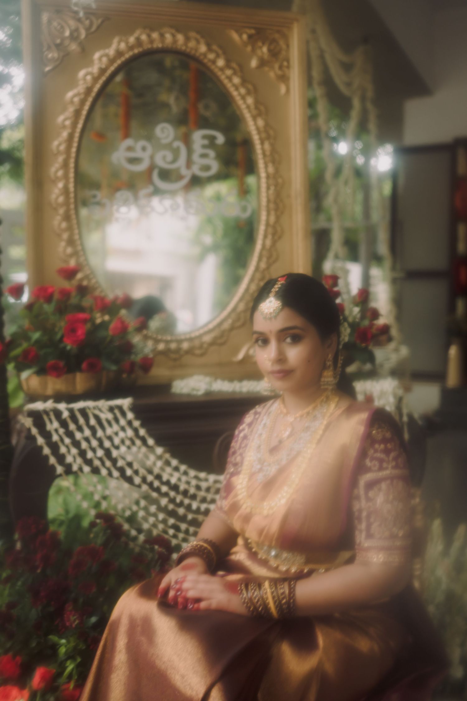 A person in traditional South Asian bridal attire poses elegantly on a sofa with red roses and ornate decor in the background.