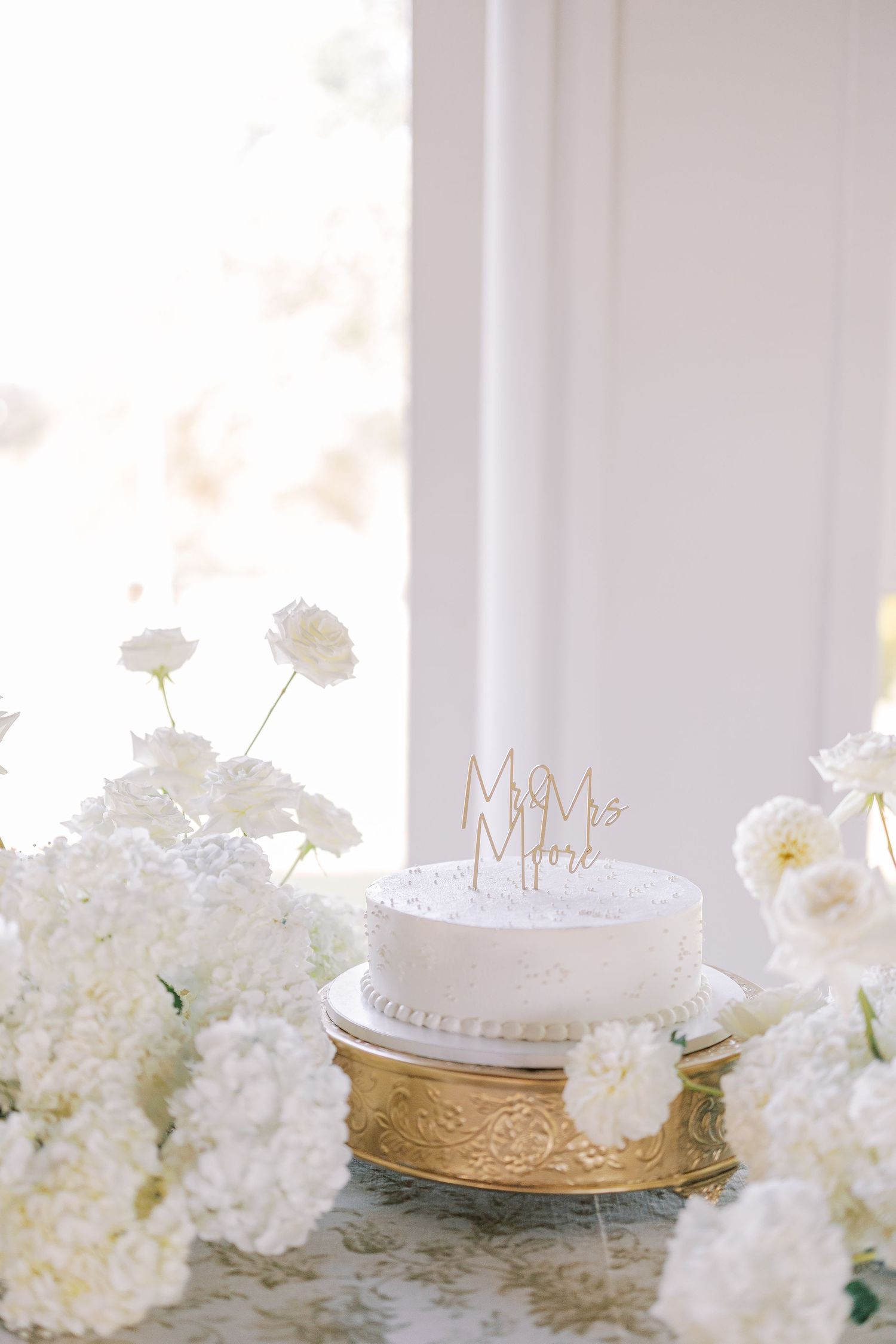 Elegant white wedding cake topped with 'Mr & Mrs' signs sits among delicate white hydrangea floral arrangements.