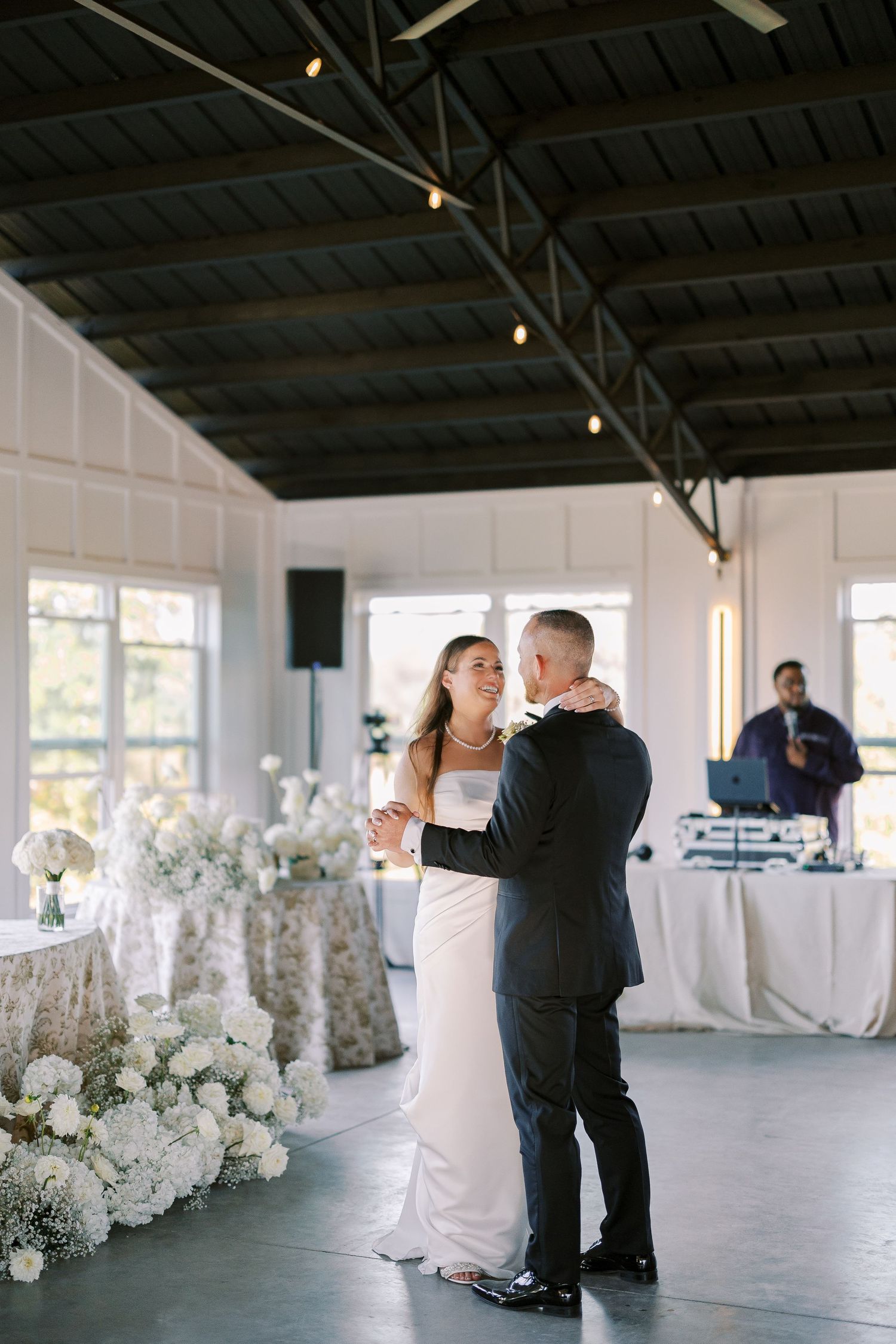 Elegant white floral arrangements line a spacious industrial-style indoor wedding venue with exposed beams and natural light.