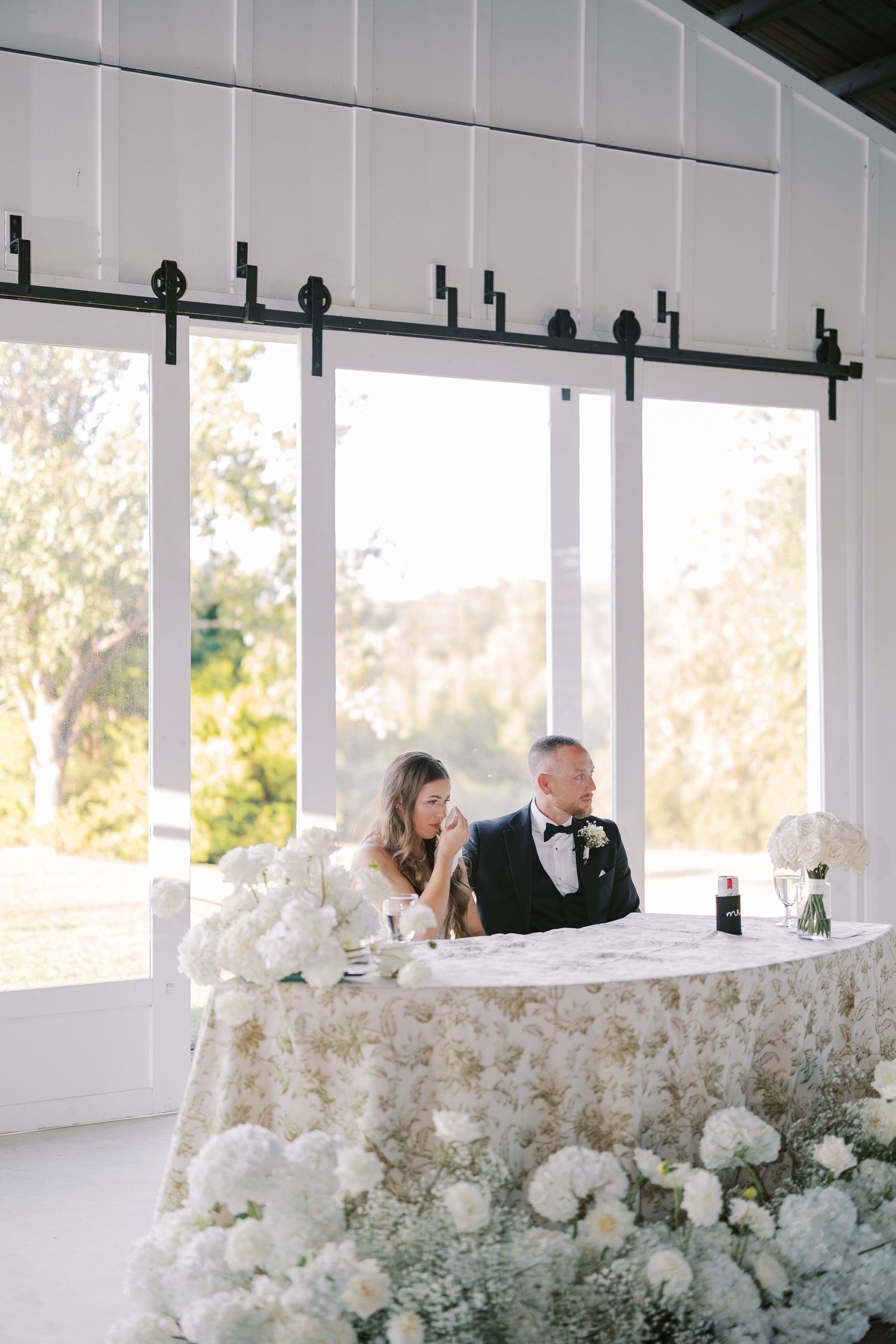 Head table at wedding reception decorated with cascading white floral arrangements and natural light from windows.