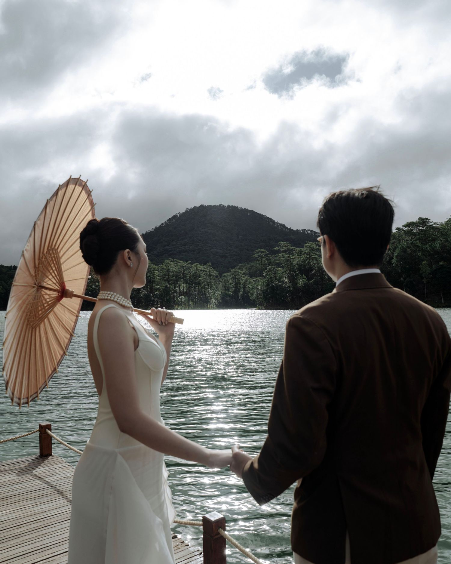 Couple holding hands on a wooden dock, bride with parasol and pearl necklace, overlooking a scenic mountain lake.