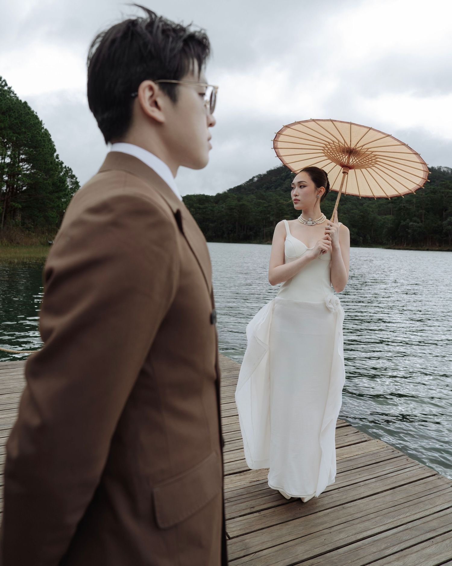 Bride holding a bamboo umbrella stands on a lake dock while groom in brown suit looks away in foreground.