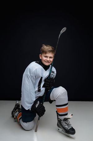 Young hockey player in white jersey and blue pants crouches in defensive position against dark background.
