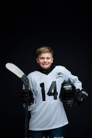 Young hockey player in a white jersey numbered 14 poses with equipment against dark background.