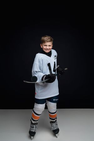 Young hockey player poses in white jersey and protective gear against dark background.